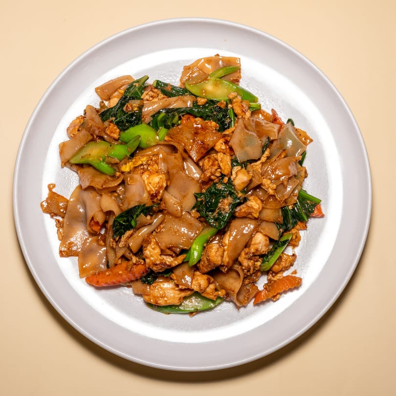 A close-up of Pad See Ew with wide noodles, chicken, Chinese broccoli, and carrots on a white plate.